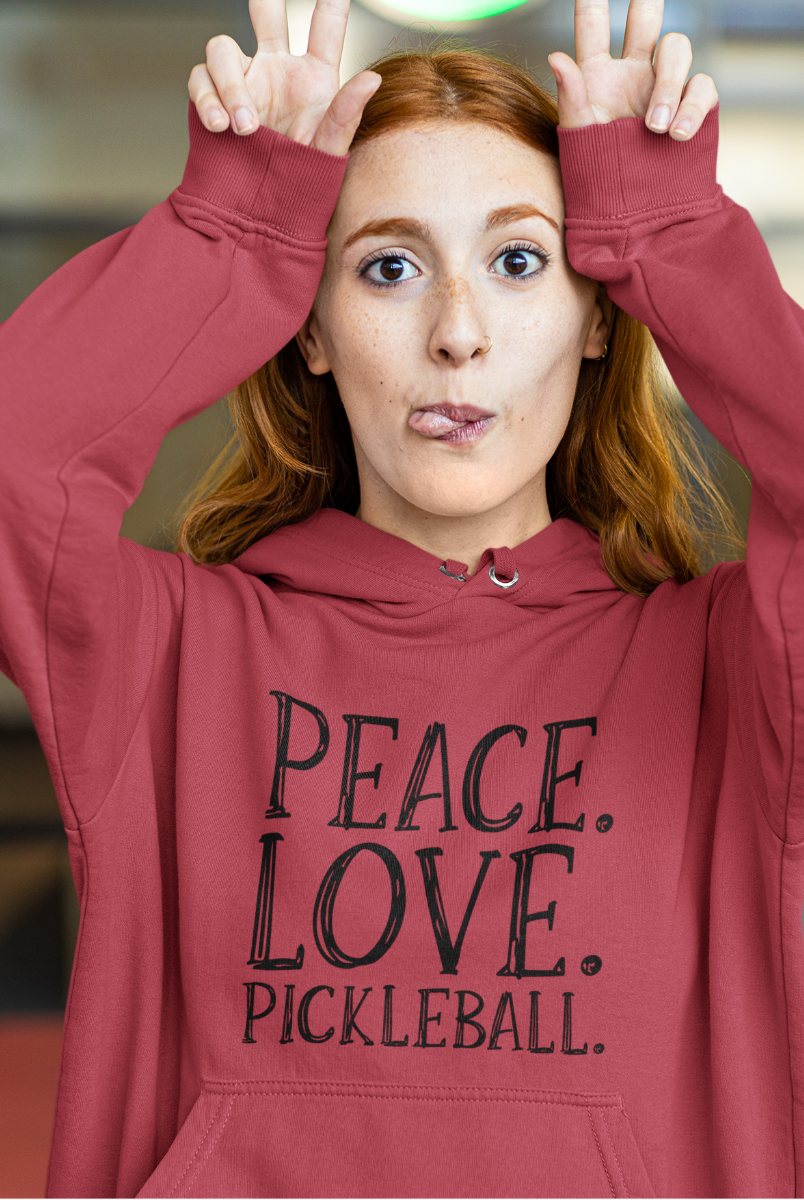 A woman with red hair wearing a dusty rose pickleball hoodie with the text 'PEACE. LOVE. Pickleball.