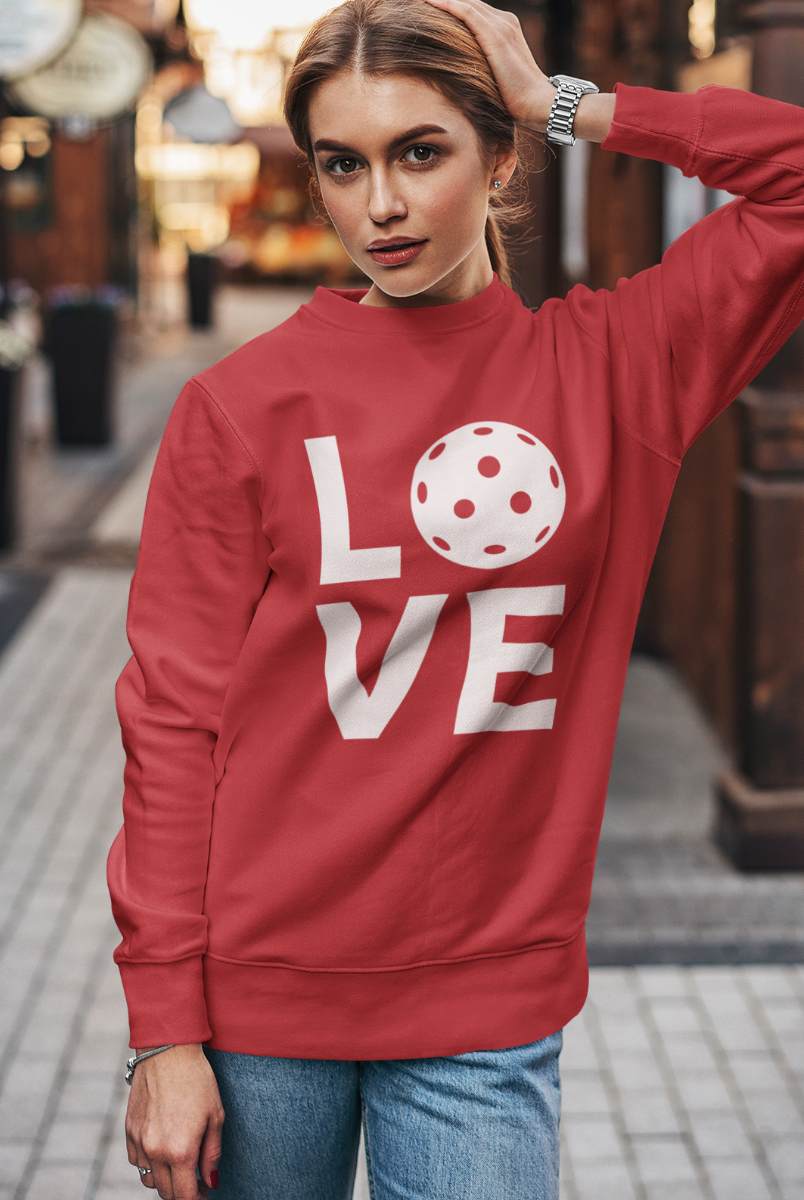 A woman wearing a red sweatshirt with a white graphic spelling 'LOVE' using a pickleball ball for the 'O', standing on a blurred street background.