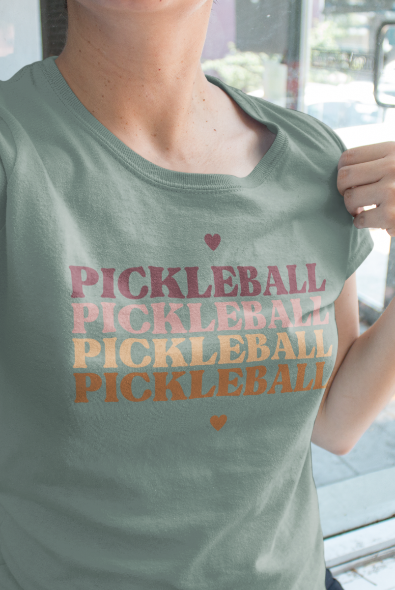 A person wearing a sage green t-shirt with 'PICKLEBALL' printed repeatedly in pink, orange, and yellow text, with small heart accents above and below.
