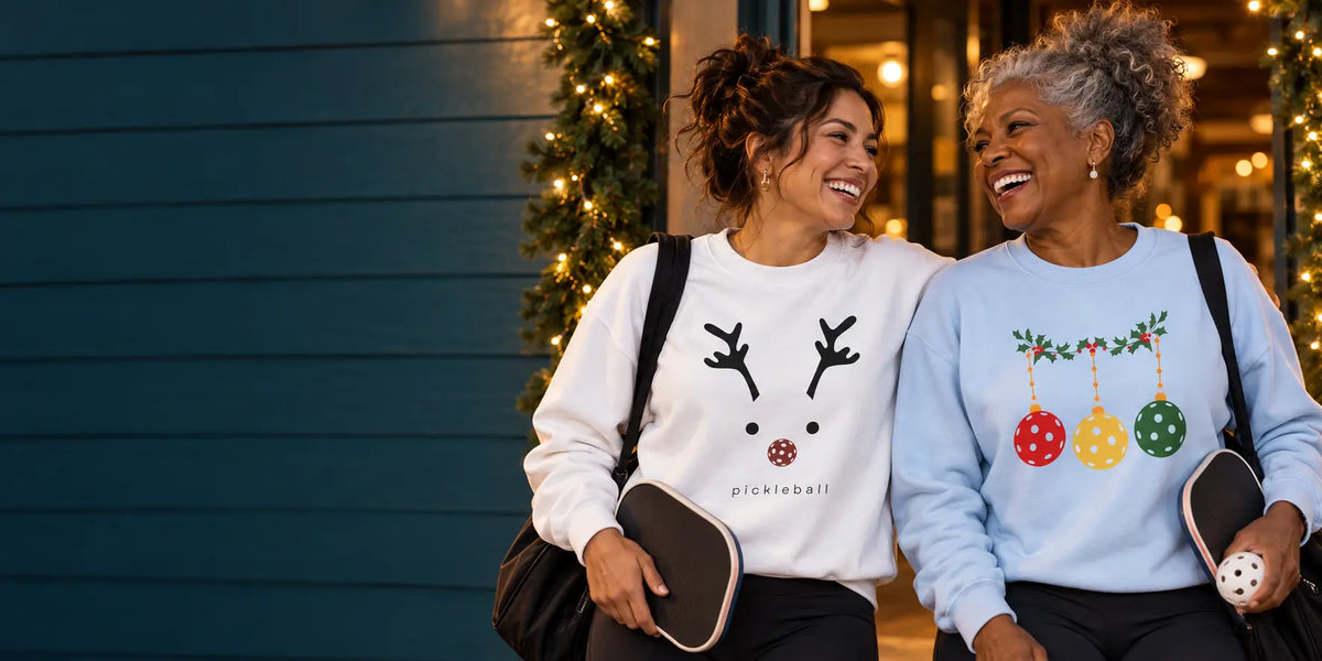 Two women wearing reindeer and pickleball ornament sweatshirts outside a festive clubhouse.