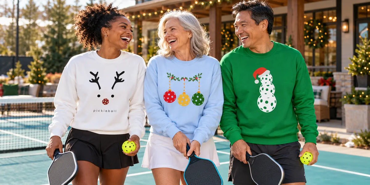 Three friends wearing pickleball Christmas sweatshirts beside an outdoor court.