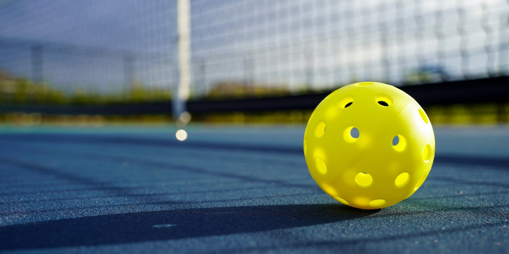 A bright yellow pickleball resting on a blue court with a net and outdoor background softly blurred.
