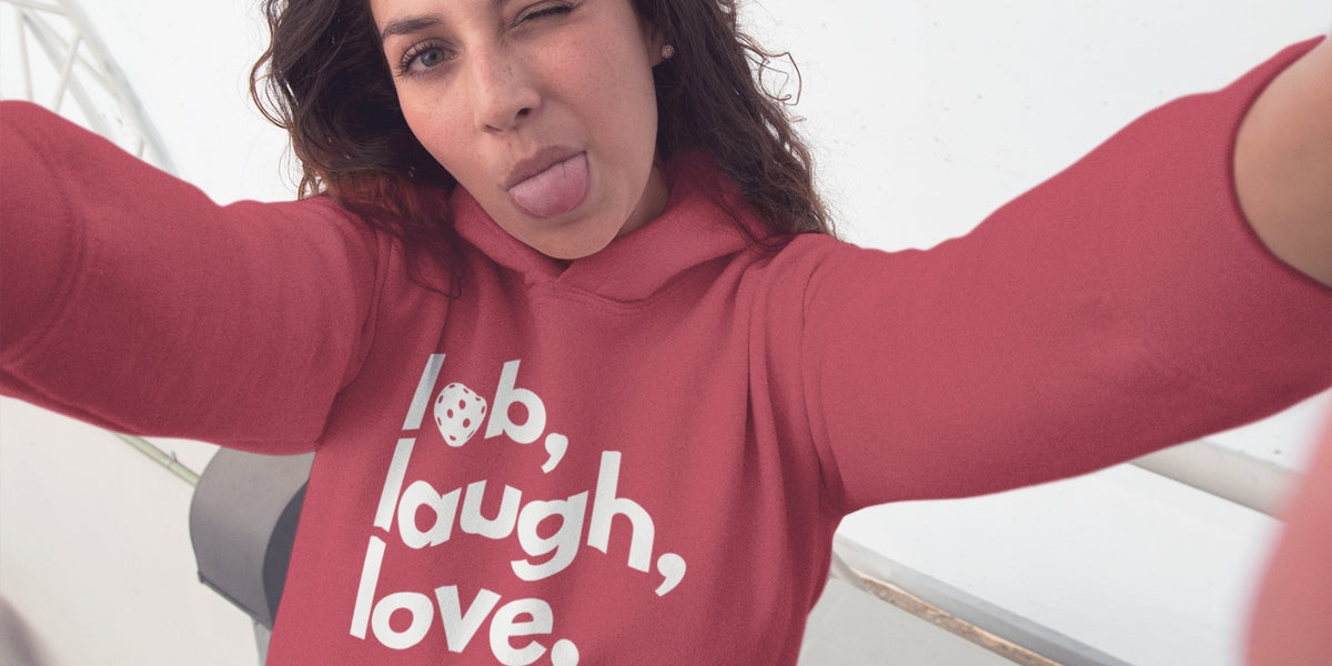 A woman wearing a dusty rose pink pickleball hoodie with the words 'lob, laugh, love.' printed in white on the front, taking a selfie with her arms outstretched.