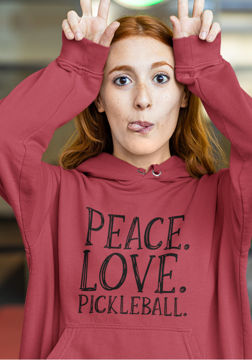 A woman with red hair wearing a dusty rose pickleball hoodie with the text 'PEACE. LOVE. Pickleball.