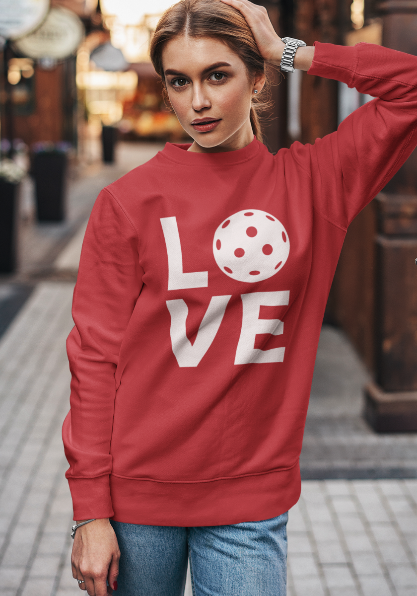 A woman wearing a red sweatshirt with a white graphic spelling 'LOVE' using a pickleball ball for the 'O', standing on a blurred street background.