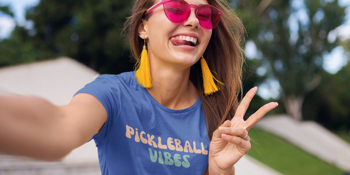 A woman taking a selfie outdoors wearing a blue graphic t-shirt that reads 'PICKLEBALL VIBES' in orange and green, along with pink sunglasses and yellow tassel earrings.