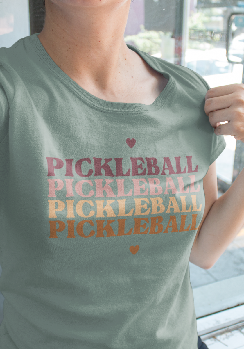 A person wearing a sage green t-shirt with 'PICKLEBALL' printed repeatedly in pink, orange, and yellow text, with small heart accents above and below.
