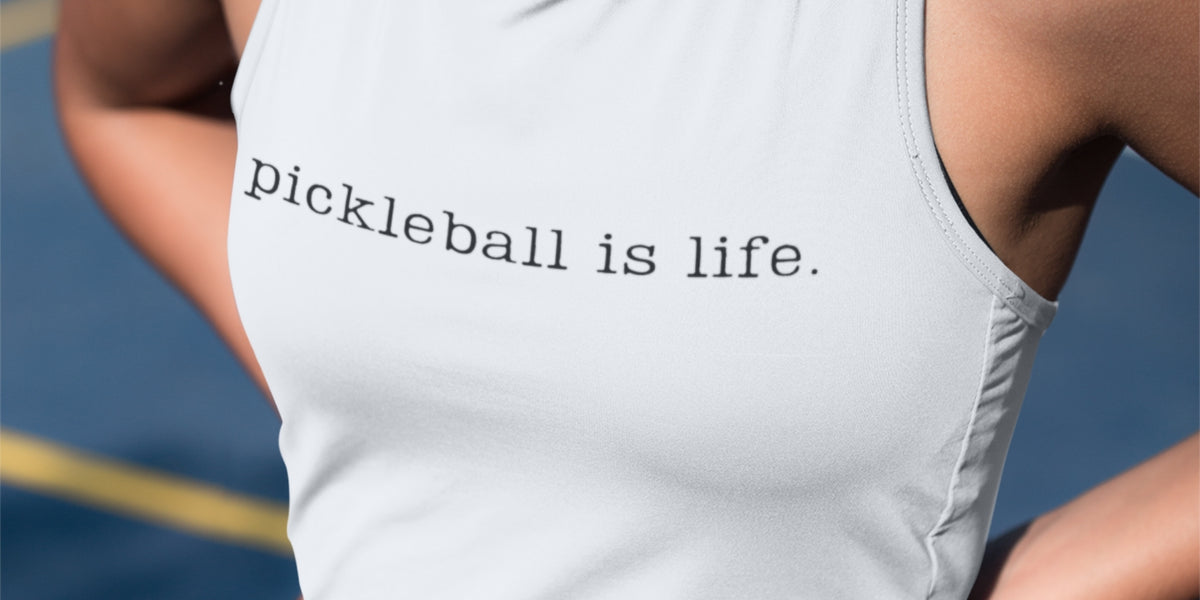 A close-up of a person wearing a white tank top with the printed phrase 'pickleball is life.' in black lowercase serif text, photographed outdoors on a blue court with a yellow line visible in the background.