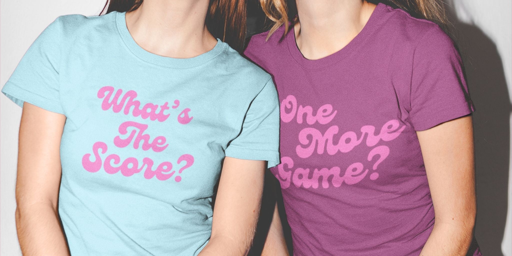 Two women wearing graphic pickleball t-shirts. The woman on the left wears a light blue t-shirt with pink text reading 'What's The Score?'.